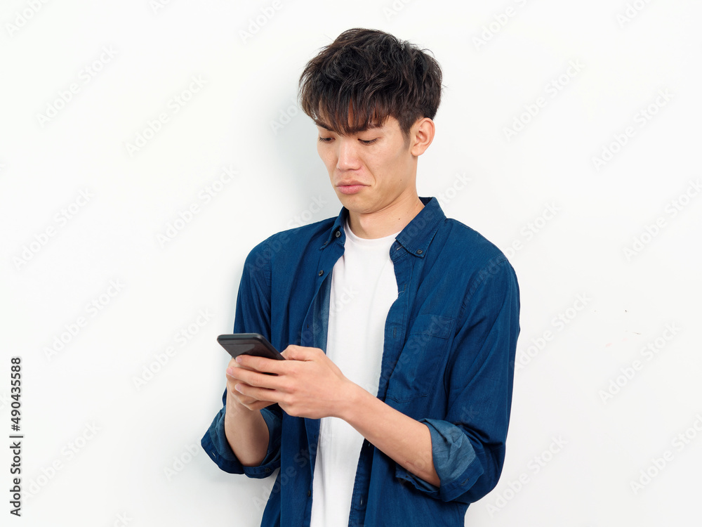 Portrait of handsome Chinese young man with curly black hair in blue shirt posing against white wall background. Looking at smartphone in both hands with disgusted expression, front view studio shot. Portrait of handsome Chinese young man with curly black hair in blue shirt posing against white wall background. Looking at smartphone in both hands with disgusted expression, front view studio shot.