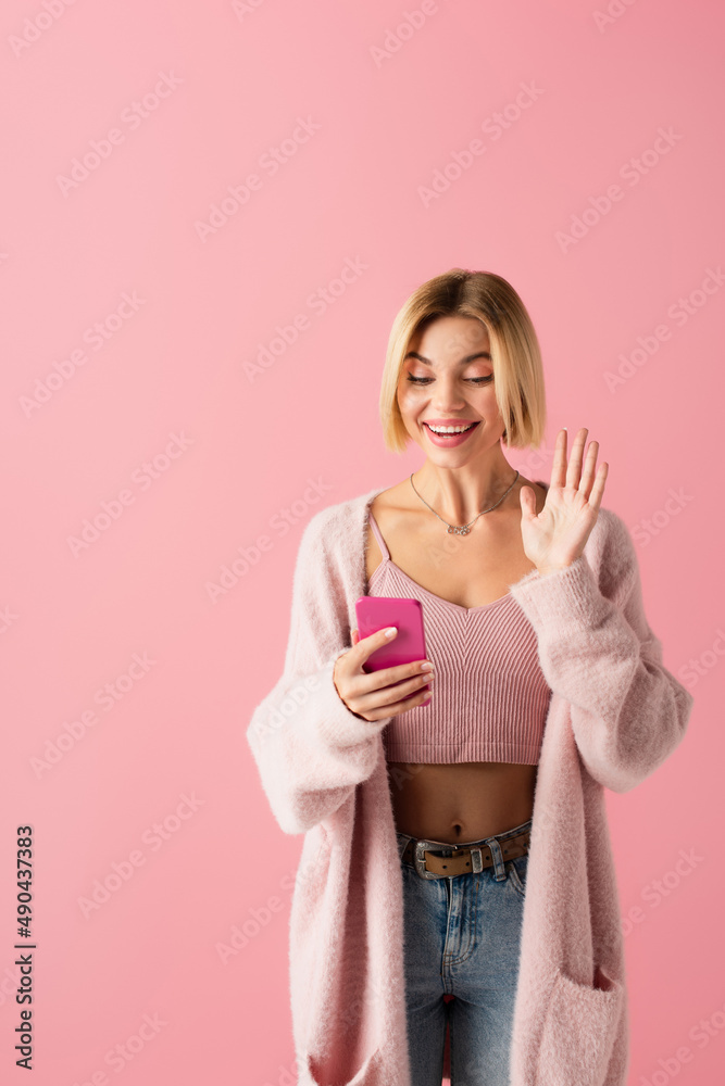 happy young woman in soft cardigan waving hand during video call isolated on pink.