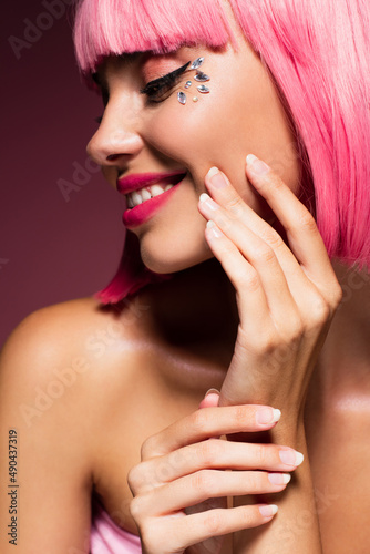 close up of cheerful woman with pink hair and shiny jewelry stones on face on dark purple.