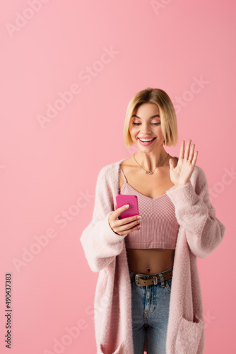 happy young woman in soft cardigan waving hand during video call isolated on pink.