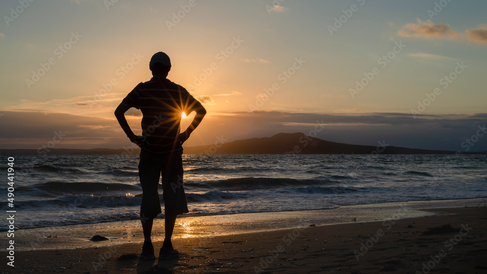Silhouette of a man watching sunrise on Milford Beach with Rangitoto Island in the distance. Auckland.