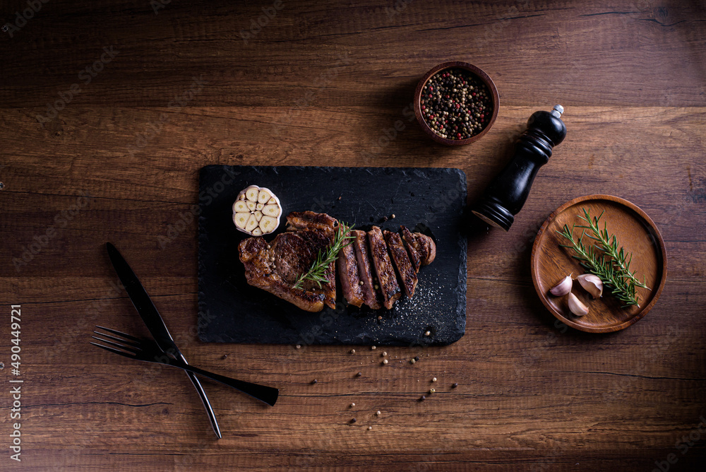Sliced striploin steak, grillid with 4seasons pepper garlic rosemary, on stone plate dark and wooden background , knife and fork black, top view Sliced striploin steak, grillid with 4seasons pepper garlic rosemary, on stone plate dark and wooden background , knife and fork black, top view