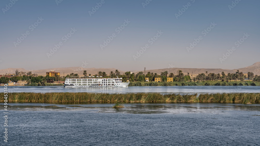 A white tourist ship sails along the shore of the Nile. Green vegetation, city houses are visible. Blue water, azure sky, sand dunes. Egypt