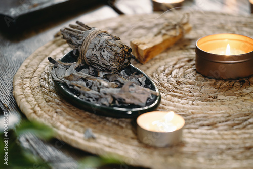 Woman hands burning white sage, before ritual on the table with candles and green plants. Smoke of smudging treats pain and stress, clear negative energy and meditation