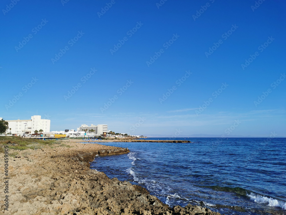 A stone shore made of solidified lava, in the distance a small sparsely populated sandy beach with sun loungers and sun umbrellas in the bay of the Mediterranean Sea on a sunny day.