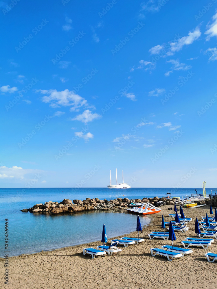 Sandy beach in a bay with sun loungers and umbrellas, in the Mediterranean Sea the largest sailing yacht in the world, an eight-deck motorsailer against a blue sky with clouds.