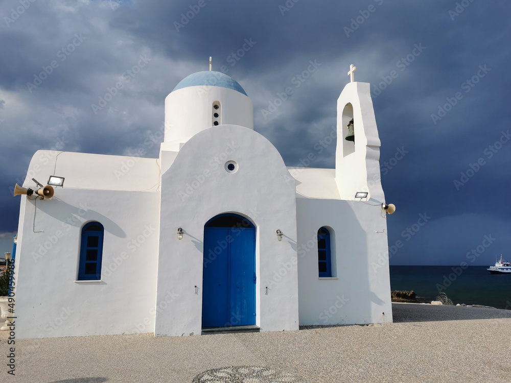 The Church of St. Nicholas the Wonderworker is white with a blue door against the backdrop of the Mediterranean Sea with a white ship and a dramatic sky.