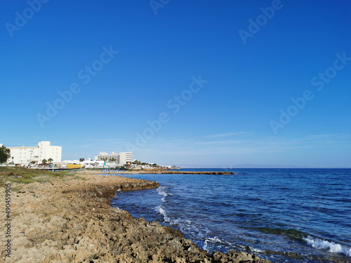 A stone shore made of solidified lava, in the distance a small sparsely populated sandy beach with sun loungers and sun umbrellas in the bay of the Mediterranean Sea on a sunny day. © Elena