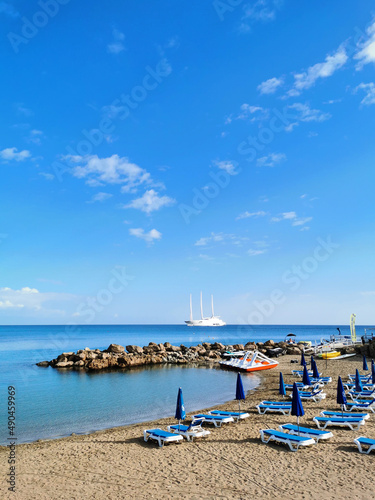Sandy beach in a bay with sun loungers and umbrellas, in the Mediterranean Sea the largest sailing yacht in the world, an eight-deck motorsailer against a blue sky with clouds. © Elena