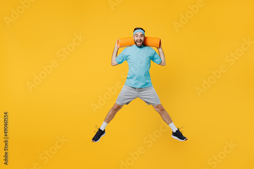 Full body overjoyed excited happy young fitness trainer instructor sporty man sportsman wear headband blue t-shirt hold yoga mat jump high isolated on plain yellow background. Workout sport concept