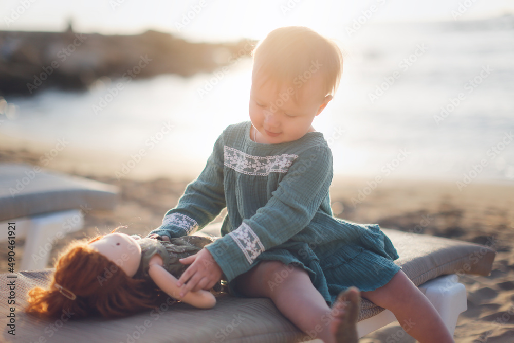 Cute european baby toddler in a green dress plays with a doll on the beach by the sea at sunset, gray background. Girl hugging waldorf soft doll, favorite toys and summer vacation kids Cute european baby toddler in a green dress plays with a doll on the beach by the sea at sunset, gray background. Girl hugging waldorf soft doll, favorite toys and summer vacation kids