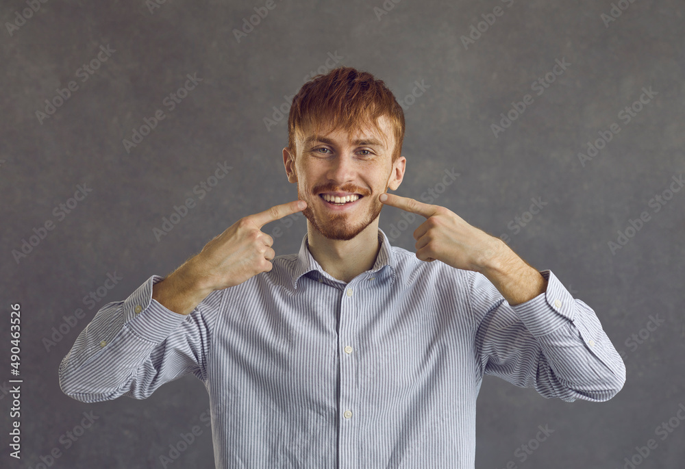 Portrait of young man wearing casual shirt showing and pointing with fingers to white teeth and mouth standing over studio grey background. Dental health, oral hygiene and stomatology concept