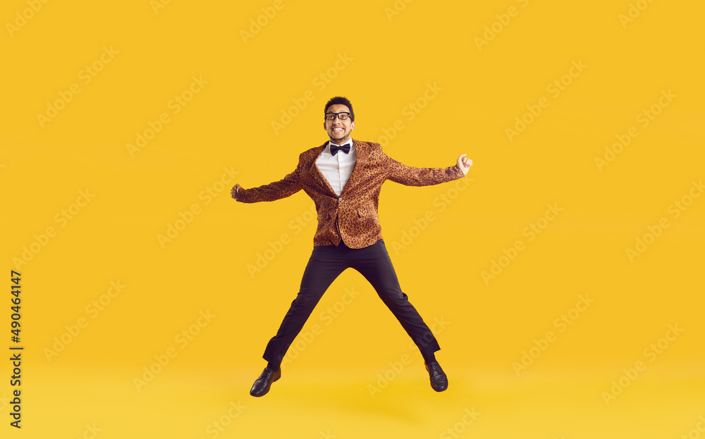 Happy dark skinned showman dressed in stylish chic having fun and celebrating his achievements. Attractive young guy in leopard jacket funny jumps isolated on yellow background. Banner. Full length.