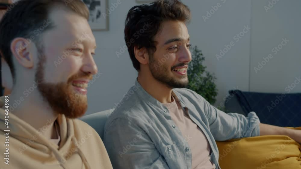 Caucasian and Middle Eastern men talking laughing drinking beer watching TV in apartment in the evening. Friendship and television concept.