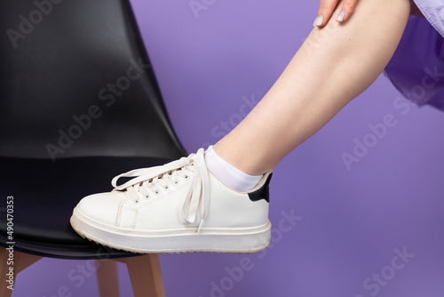 Cropped photo of a young woman leg in white sneekers putting on a chair on purple background, health slim legs concept.