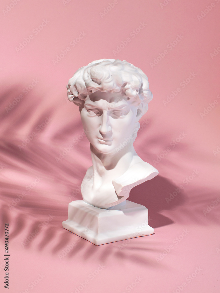 The plaster head of a handsome man against pastel pink background, the shadow of a palm branch over it. Summer tropical mood. The plaster head of a handsome man against pastel pink background, the shadow of a palm branch over it. Summer tropical mood.
