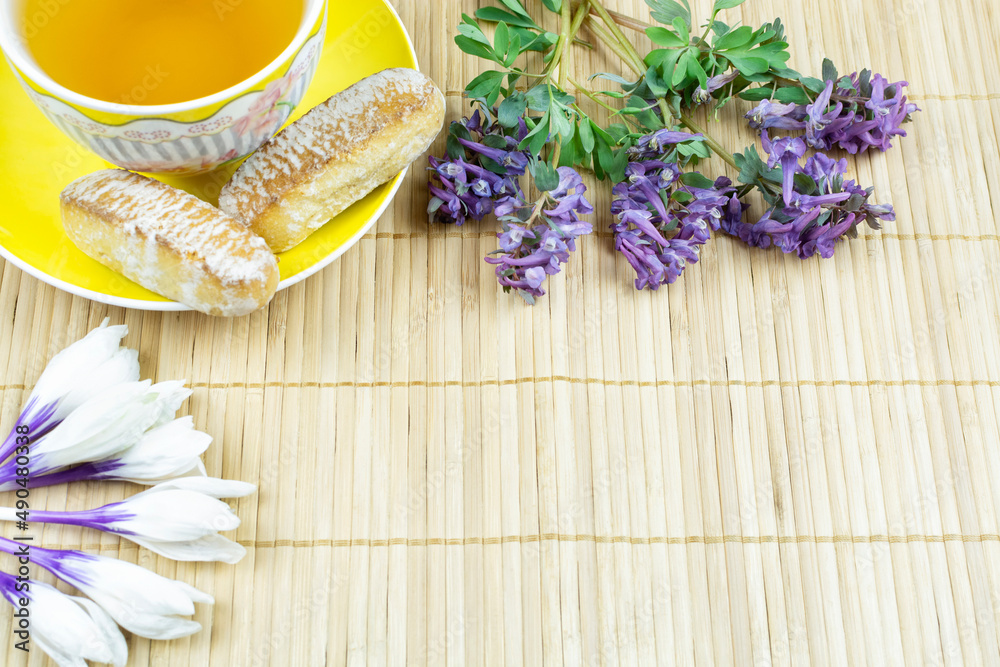 Good morning. Yellow Cup of tea on a saucer, gingerbread, spring flowers crocuses and Corydalis on a bamboo background. Space for text. The concept of the celebration.