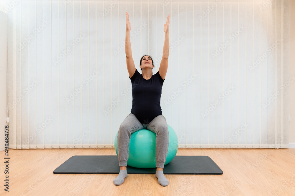 Fototapeta premium A pregnant woman doing pilates exercises sitting on a fitness ball with her arms stretched upward and looking up