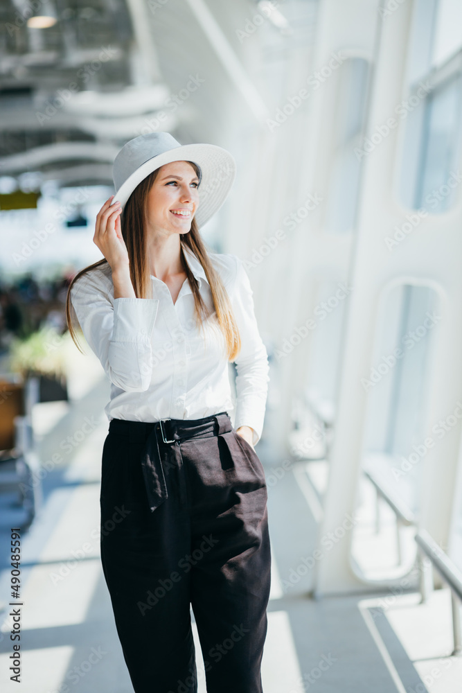 Fototapeta premium Cool young woman in boater and white dress smiles and looks into camera outside. Tourist holds passport and poses on crosswalk near airport.