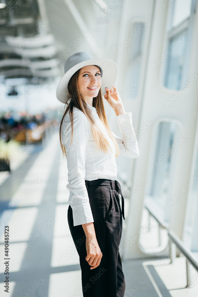 Fototapeta premium Airport business woman on smart phone at gate waiting in terminal. Air travel concept with young casual businesswoman sitting with hand luggage suitcase. Beautiful young female professional
