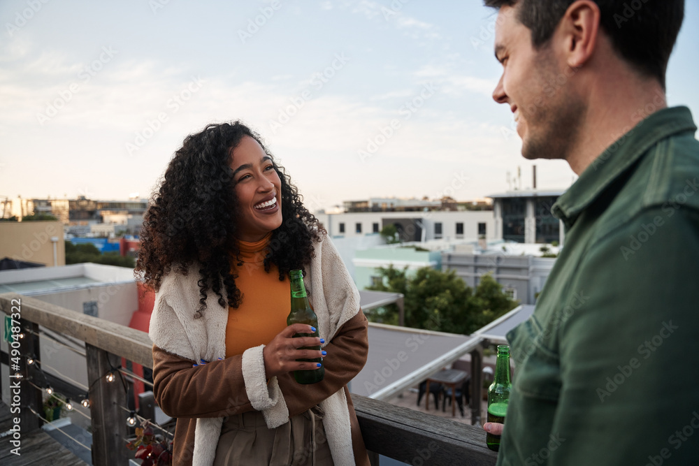 Fototapeta premium Biracial young adult female laughing with a friend on the balcony of a rooftop terrace in the city. Drink in hand, cityscape views