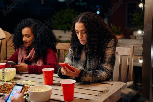 Two biracial young adult females sitting at a party, texting on her phone. Anti-social, using technology at rooftop party
