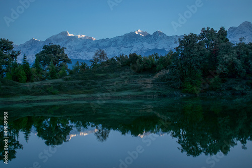 Snow covered mountains surrounded by forest & clear blue skies perfectly reflected in lake on a clear morning. Amazing Himalaya landscape during spring trek to Deoria lake, Chopta, Uttarakhand (India)