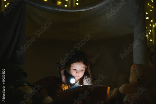 child girl reading with book and flashlight and teddy bear in tent. before going to bed