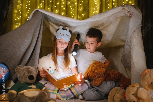 reading and family games in children's tent. boy and girl with book and flashlight before going to bed © Serhii