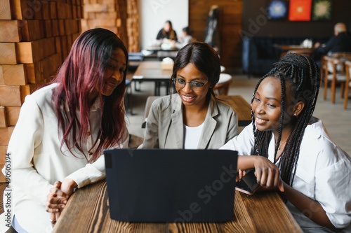 Three African American girls (students) sitting at the table in cafe studying up for test or making homework together, they are using laptop and digital tablet.