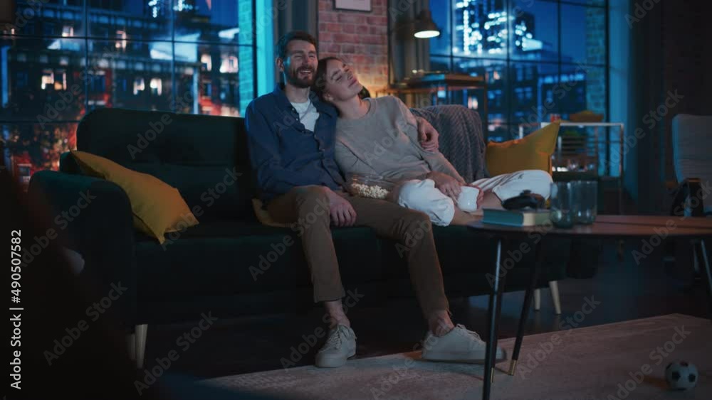 Portrait of Beautiful Couple Spending Time at Home, Sitting on a Couch, Hugging and Watching Exciting TV Show in Their Stylish Loft Apartment. Man and Woman Streaming Comedy Movie and Have Good Time. Portrait of Beautiful Couple Spending Time at Home, Sitting on a Couch, Hugging and Watching Exciting TV Show in Their Stylish Loft Apartment. Man and Woman Streaming Comedy Movie and Have Good Time.
