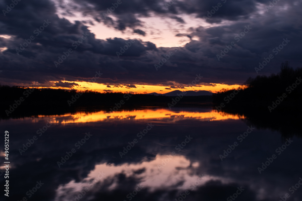 Twilight landscape with dark ominous storm clouds and fading orange glow at sky above horizon - symmetric reflection
