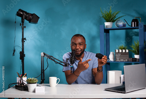 Vlogger recording npf rechargeable battery pack review for pointing out advantages talking into studio microphone. Influencer talking about lithium ion accumulator sitting at desk. photo