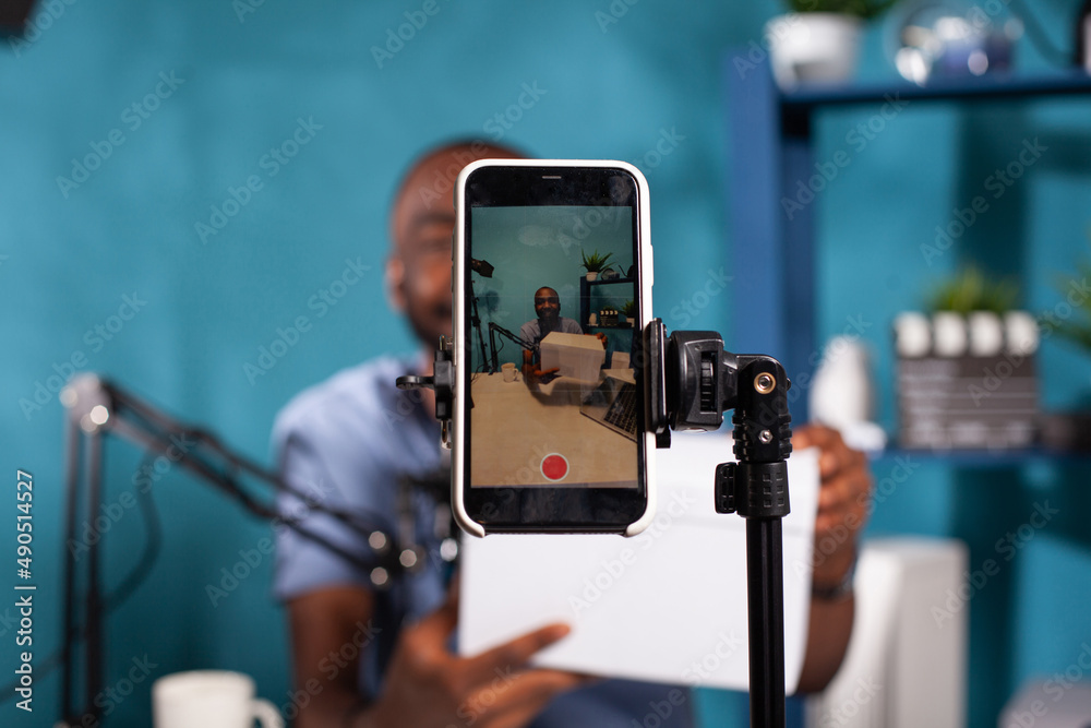 Closeup of live vlog setup with smarthone on stand filming smiling vlogger hosting online giveaway holding product box in vlogging studio. Selective focus on mobile phone recording influencer.