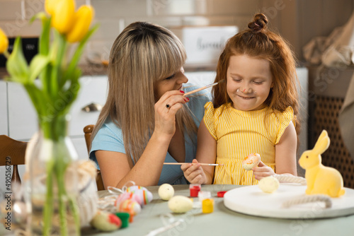 Playful mother and daughter sitting at the kitchen table and painting eggs for Easter