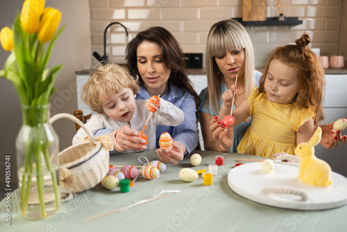 Two women and their children are sitting in the kitchen at the table and painting Easter eggs