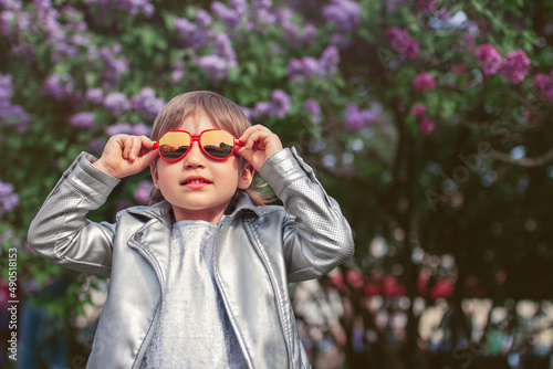 little girl in red sunglasses in silver jacket smiles and walks in flowering park in spring on the background of flowering trees © ksenija1803z