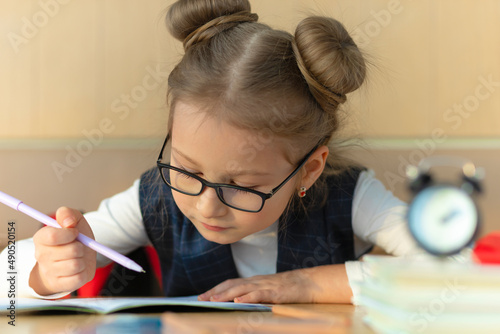 little schoolgirl girl in glasses doing homework, studying at school. alarm clock and lunch break. concept of child education, deadline, poor eyesight