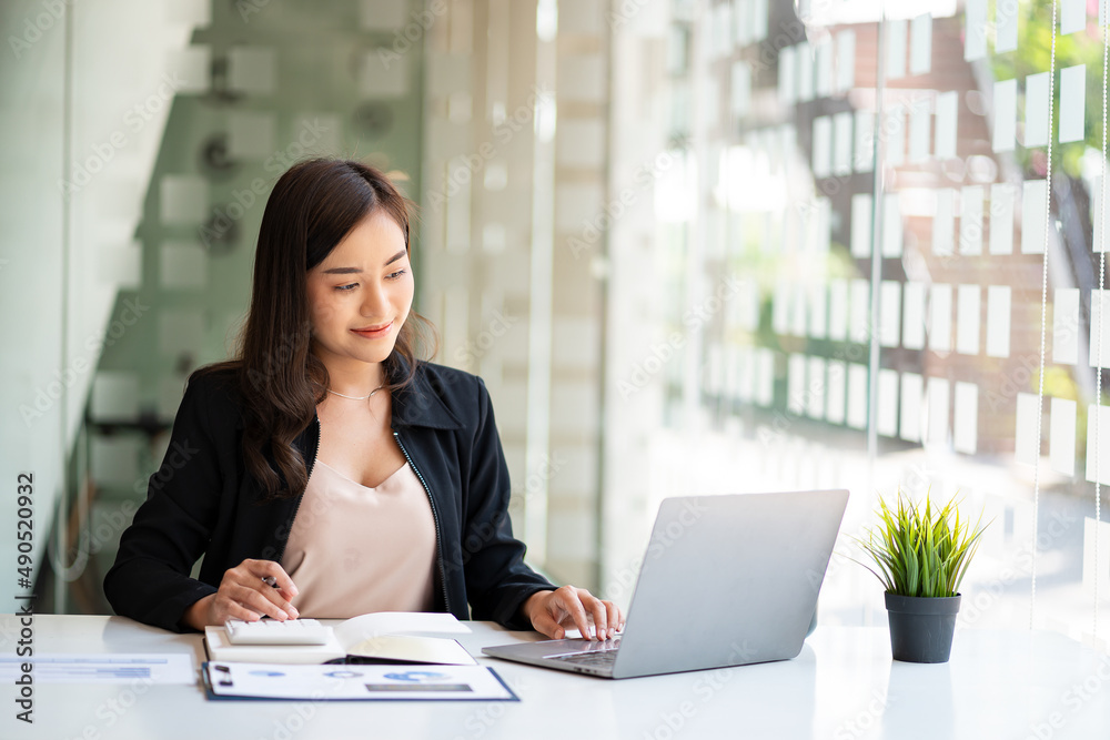 Asian businesswoman working in financial accounting analyzes a chart using a laptop calculator with documents and holding a pen sitting on a chair at an office desk. Asian businesswoman working in financial accounting analyzes a chart using a laptop calculator with documents and holding a pen sitting on a chair at an office desk.