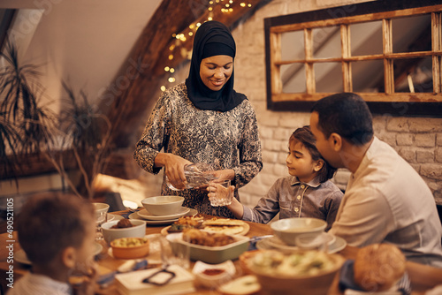 Happy Muslim mother pours water into daughter s glass during family meal at home.