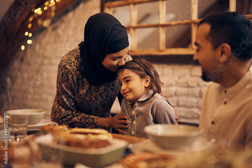 Loving Muslim mother kisses her daughter during family meal at dining table.