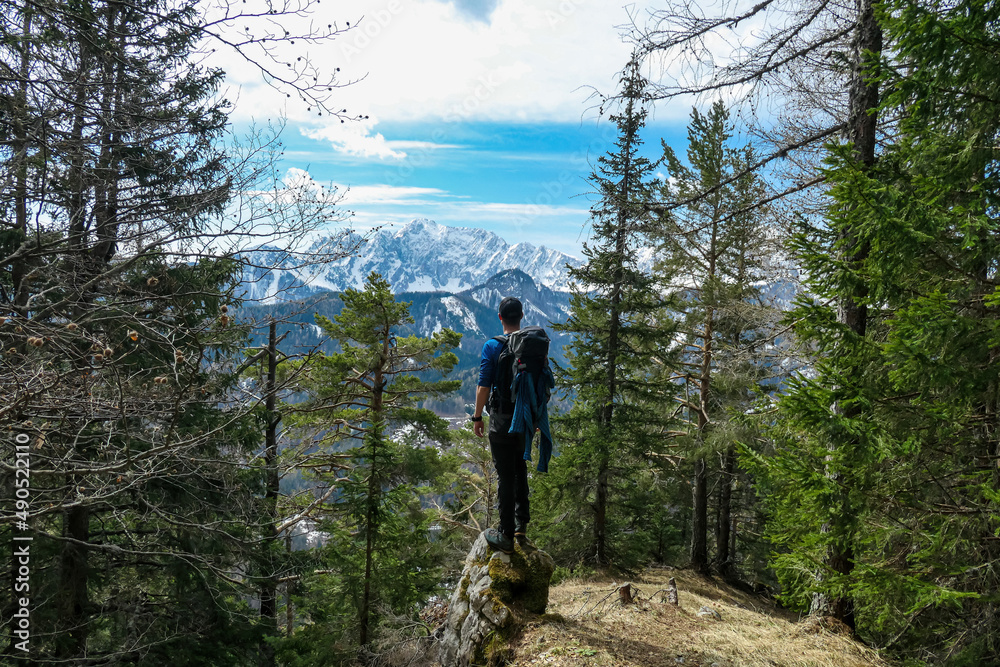 Man with backpack standing on a rock with scenic view of snow capped mountain peaks of Karawanks near Sinacher Gupf in Carinthia, Austria. Mount Wertatscha is visible through dense forest in spring