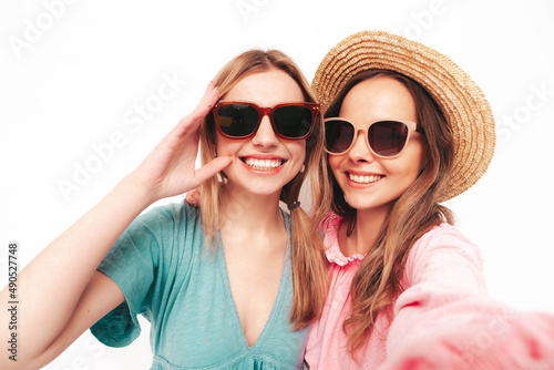 Two young beautiful smiling brunette hipster female in trendy summer dresses. Sexy carefree women posing near pink wall. Positive models having fun.Taking Pov selfie. In hats and sunglasses