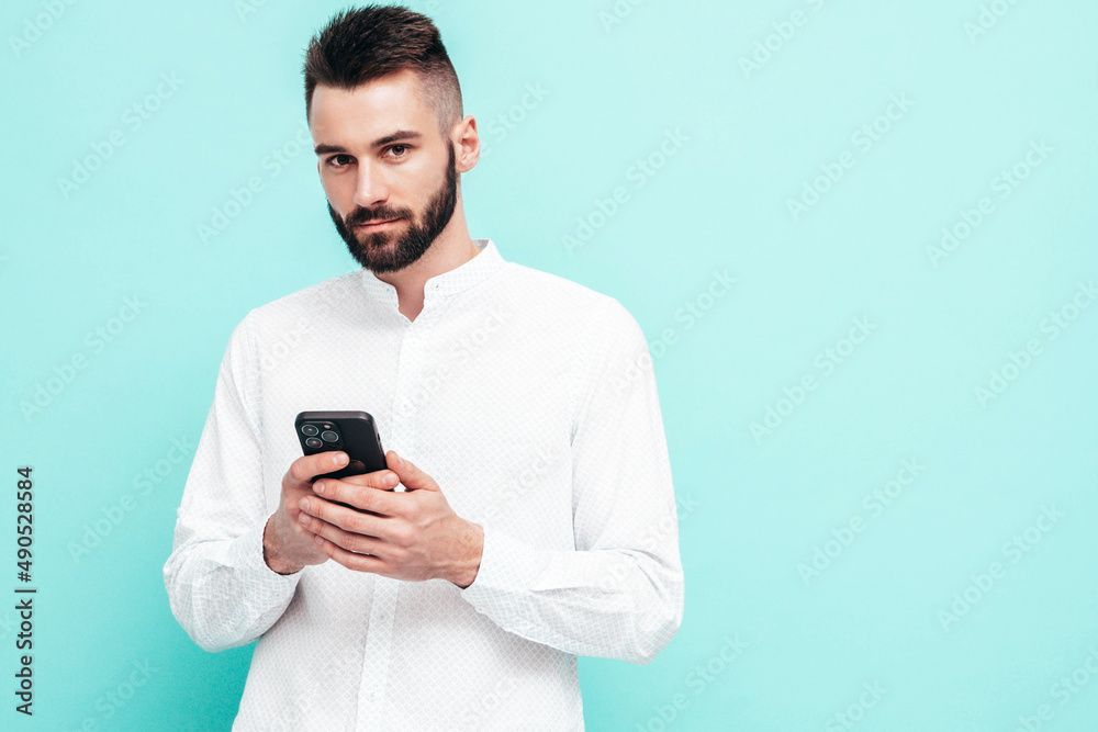 Handsome confident model.Sexy stylish man dressed in shirt and jeans. Fashion hipster male posing near blue wall in studio. Holding smartphone. Looking at cellphone screen. Using apps Handsome confident model.Sexy stylish man dressed in shirt and jeans. Fashion hipster male posing near blue wall in studio. Holding smartphone. Looking at cellphone screen. Using apps