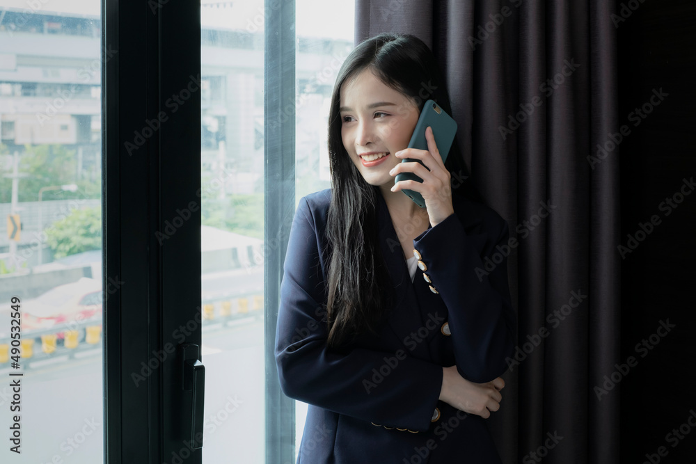 Young businesswoman using smartphone working in hotel room during her business travel. Businesswoman working from hotel room on business trip standing by the window in sunny day. Young businesswoman using smartphone working in hotel room during her business travel. Businesswoman working from hotel room on business trip standing by the window in sunny day.