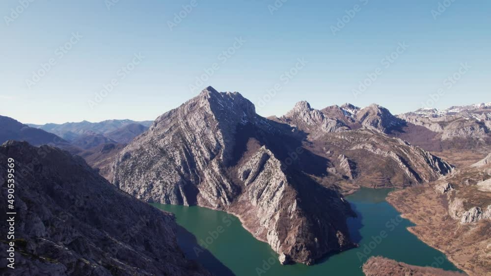 Greenish river split in two by a huge rocky brown mountain in León, Spain. Mountain range below a clear, blue sky in plain daylight. Drone aerial shot, forward motion. Greenish river split in two by a huge rocky brown mountain in León, Spain. Mountain range below a clear, blue sky in plain daylight. Drone aerial shot, forward motion.