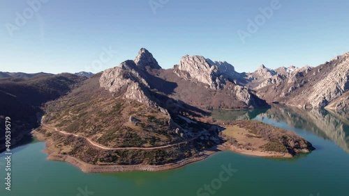 Sharp, tall mountain range bext to a greenish water lake in León, Spain. Rocky mountains reflected on resrervoir surface. Calm swamp next to mountain foot. Aerial drone shot moving forward.    photo