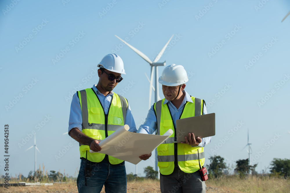 two architects working on a site. two Asian male engineers working on site with wind turbine propeller and clear blue sky on the background. Alternative energy, environmental friendly for the future. two architects working on a site. two Asian male engineers working on site with wind turbine propeller and clear blue sky on the background. Alternative energy, environmental friendly for the future.