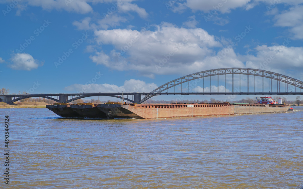 Obraz premium Nijmegen, Netherlands - February 27. 2022: View over river waal with inland waterway ship and arch bridge on two pylons against blue sky
