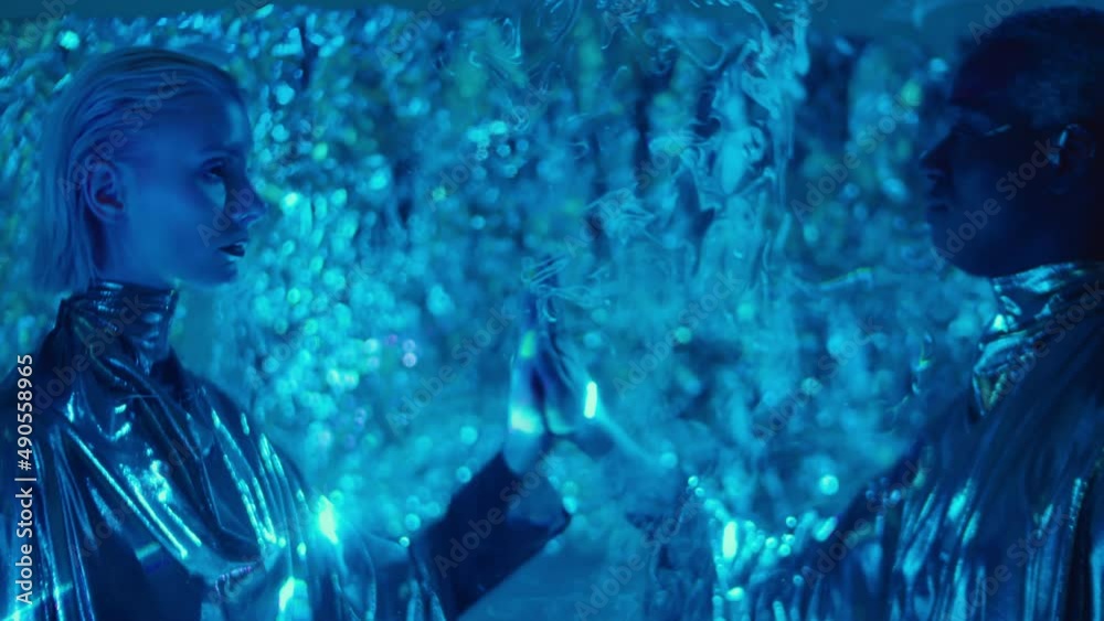 Cosmic man and woman in silver outfits standing in dark room on both sides of flat sheet of liquid sky laser light and touching hands of each other